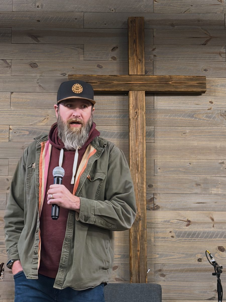 Michael Suntribo preaching with a microphone in front of a large wooden cross on a wood-plank wall